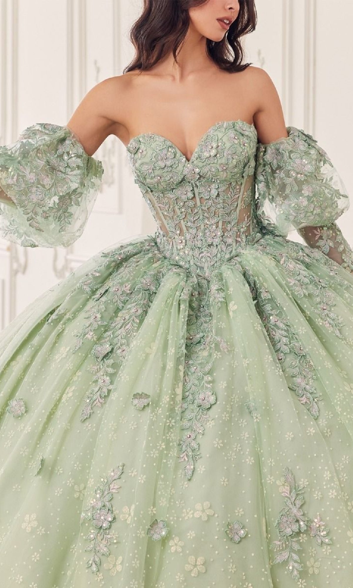 A woman wears the Quinceanera Dress 15722 by Ladivine—a strapless, pastel green ball gown with floral embroidery and removable puff sleeves—set against a light, ornate background.